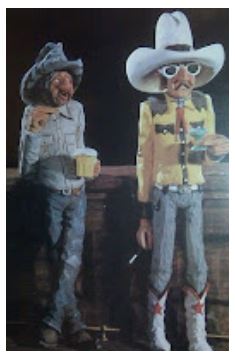 Photo of two comical cowboy statues holding drinks at a bar. The statues appear to be carved from wood. The statue on the left is an old, rough cowboy wearing simple working cowboy clothes and holding a mug of beer. The cowboy on the right holds a martini and wears fancy clothes, boots, and sunglasses. 